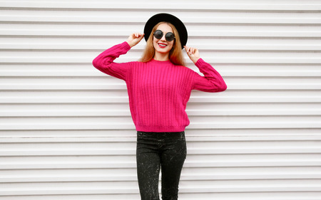 Stylish happy young woman in colorful pink knitted sweater, black round hat posing on city white wall backgroundの写真素材
