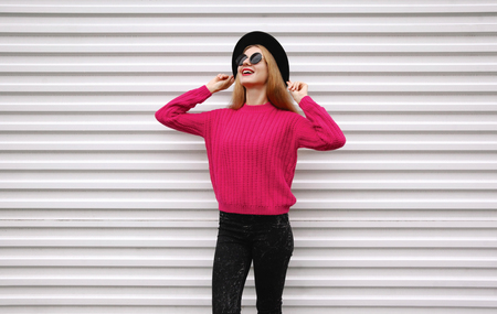 Stylish smiling young girl in colorful pink knitted sweater, black round hat on white wall backgroundの写真素材