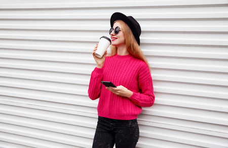 Stylish young smiling woman drinks coffee holds phone in pink knitted sweater on white wall backgroundの写真素材