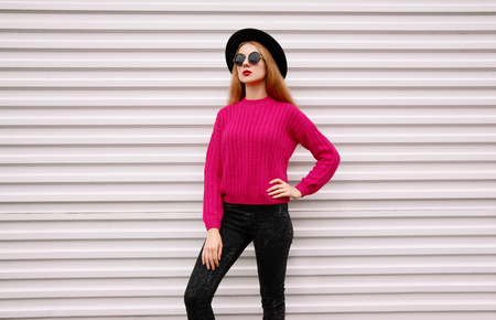 Stylish young woman posing in colorful pink knitted sweater, black round hat on white wall backgroundの写真素材