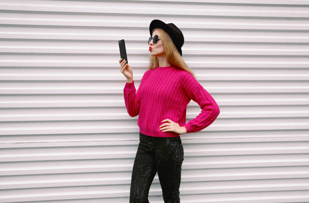 Fashion, technology and people concept - woman taking selfie picture by smartphone blowing red lips sending sweet air kiss in colorful pink knitted sweater, black round hat on white wall background on city streetの写真素材