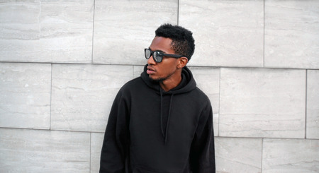 Portrait african man in black hoodie, sunglasses looking away on city street over gray brick wall backgroundの写真素材