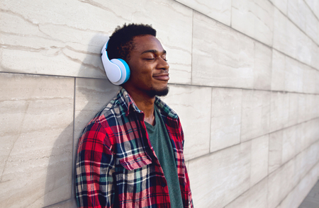 Portrait happy smiling african man in wireless headphones enjoying listening to music on city street over gray wall backgroundの写真素材