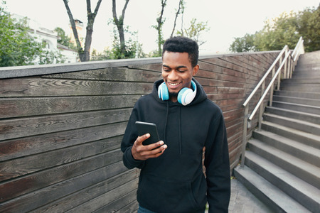 Portrait happy smiling african man with phone, headphones listening to music wearing black hoodie, walking on city streetの写真素材