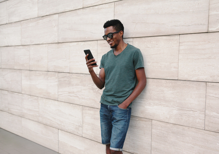 Portrait happy smiling african man with phone in green t-shirt, sunglasses on city street over gray brick wall backgroundの写真素材
