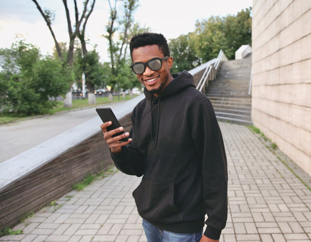 Portrait happy smiling african man with phone in black hoodie, sunglasses on city streetの写真素材