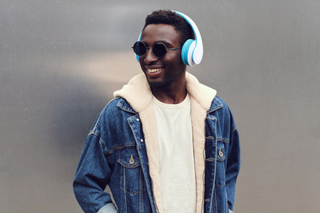 Portrait stylish urban smiling african man in headphones enjoying listening to music on gray metal wall background, wearing jeans jacketの写真素材