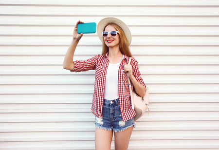 Happy smiling woman taking selfie picture by phone in summer round straw hat, checkered shirt, shorts on white wall backgroundの写真素材