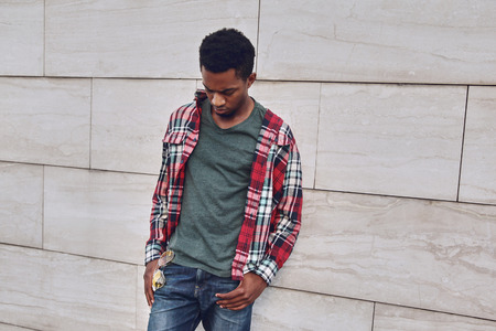 Stylish african man wearing red plaid shirt looking down, young guy posing on city street, gray brick wall backgroundの写真素材