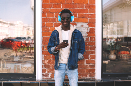 Stylish cheerful african man using smartphone listening to music in wireless headphones on city street over brick wall backgroundの写真素材