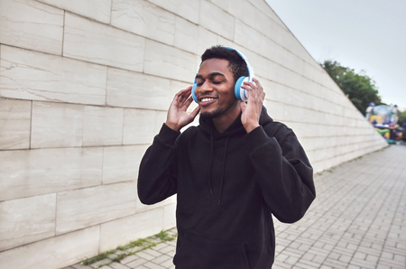 urban happy smiling african man in wireless headphones enjoying listening to music wearing black hoodie on city street over gray wall backgroundの写真素材