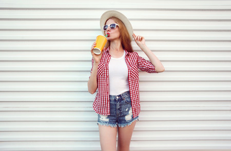 Happy woman enjoying fresh orange juice in summer round straw hat, checkered shirt, shorts on white wall backgroundの写真素材