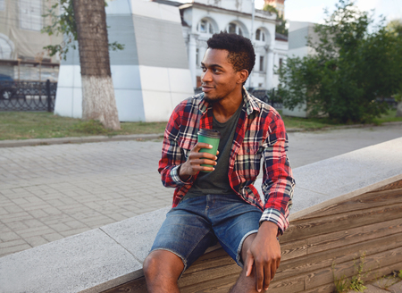 Break time! Young african man with coffee cup sitting on city streetの写真素材