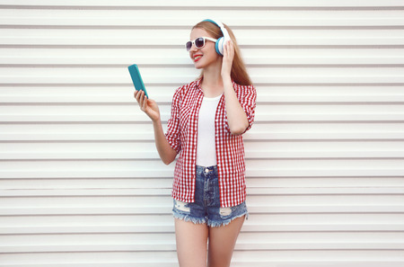 Happy smiling young woman in headphones with smartphone listening to music wearing checkered shirt, shorts on white wall backgroundの写真素材
