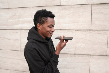 portrait smiling african man holding in hands smartphone using voice command recorder, assistant or calling, looking at phone walking on city street on gray wall backgroundの写真素材
