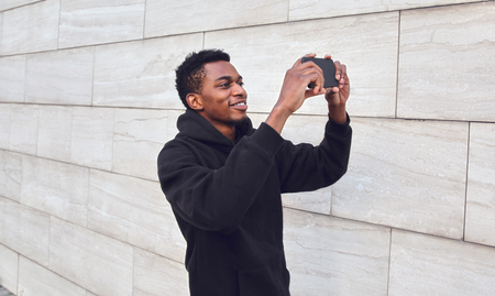 Cheerful smiling african man taking selfie picture by phone on city street over gray wall backgroundの写真素材