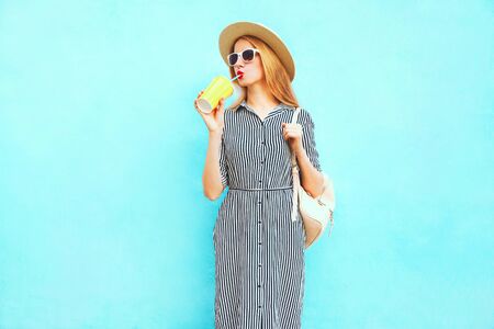 Fashion woman drinking fruit juice from cup, wearing summer round straw hat, striped dress on blue backgroundの写真素材