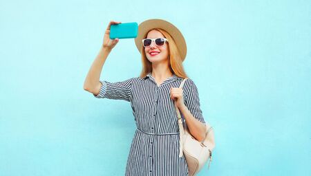 Happy smiling woman taking selfie picture by phone in summer round straw hat, striped dress with backpack on blue backgroundの写真素材
