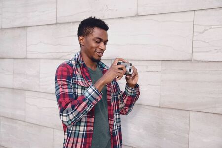 Portrait happy smiling young african man with vintage film camera taking picture walking on city street over gray brick wall backgroundの写真素材