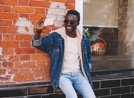 Happy smiling african man taking selfie picture by smartphone sitting on city street over brick textured wall backgroundの写真素材