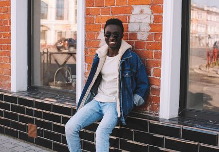 Stylish happy smiling african man wearing jeans jacket sitting on city street over brick wall backgroundの写真素材