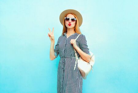Cool girl with red lips sending sweet air kiss in summer round straw hat with backpack on blue backgroundの写真素材