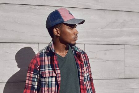 Portrait close up stylish african man looking away wearing baseball cap, red plaid shirt, young guy posing on city street over gray brick wall backgroundの写真素材