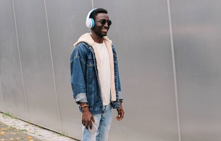 Cool modern smiling african man in wireless headphones listening to music on phone while walking on city street over gray metal wall backgroundの写真素材