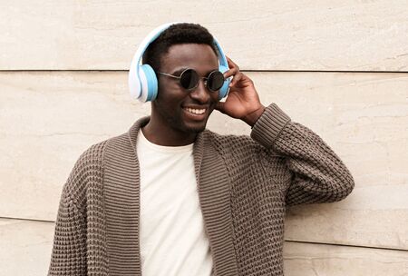 Portrait stylish smiling african man in wireless headphones listening to music wearing brown knitted cardigan and sunglasses on city street over brick wall backgroundの写真素材