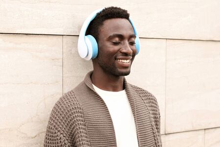 Portrait stylish smiling african man in wireless headphones listening to music wearing brown knitted cardigan and sunglasses on city street over brick wall backgroundの写真素材