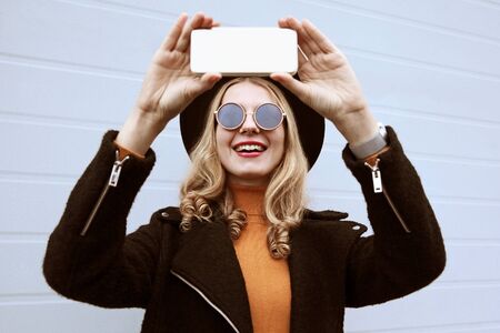 Portrait stylish happy smiling woman taking selfie picture by smartphone wearing black coat and hat on city street over wall backgroundの写真素材