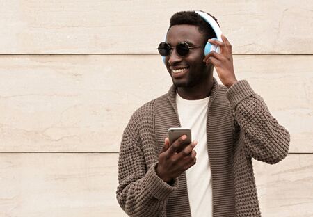 Portrait stylish smiling african man with phone in wireless headphones listening to music wearing brown knitted cardigan and sunglasses on city street over brick wall backgroundの写真素材