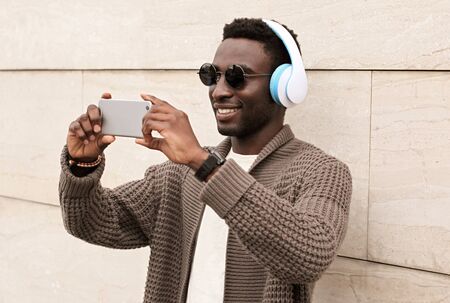 Portrait stylish smiling african man taking selfie picture by smartphone in wireless headphones listening to music wearing brown knitted cardigan, sunglasses on city street over wall backgroundの写真素材