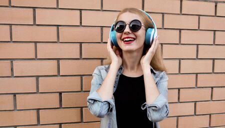 Portrait of happy smiling woman in wireless headphones listening to music wearing a black round hat, jeans jacket over brick wall backgroundの写真素材