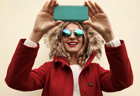 Portrait happy smiling young woman taking selfie picture by smartphone wearing red jacket with fur...