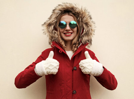 Portrait happy smiling young woman showing thumbs up wearing red jacket with fur hood and white...