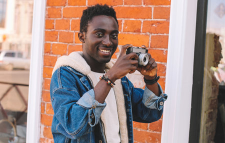 Portrait of happy smiling african man photographer with vintage film camera taking picture walking on city streetの写真素材