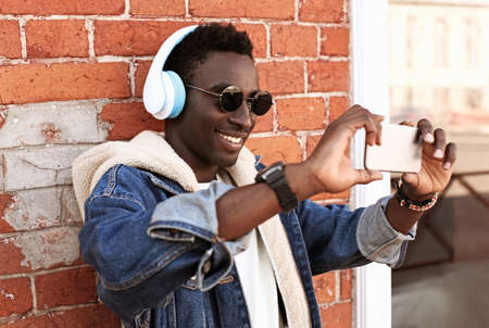 Portrait of happy smiling young african man taking selfie by smartphone listening to music in headphones on a city streetの写真素材
