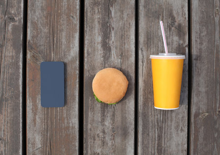Fast food, unhealthy diet concept - phone with juice cup and burger on a wooden table or floor backgroundの写真素材