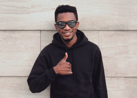 Portrait close up of happy smiling young african man showing thumbs up wearing a black hoodie, sunglasses on a city street over gray wall backgroundの写真素材