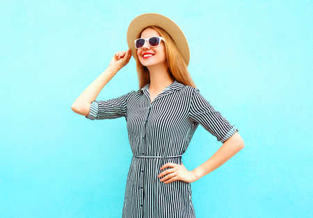 Portrait of happy smiling young woman wearing a summer straw hat, striped dress on a blue backgroundの写真素材