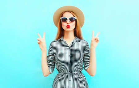 Portrait of beautiful young woman blowing her red lips wearing a striped dress, summer straw hat on a blue backgroundの写真素材