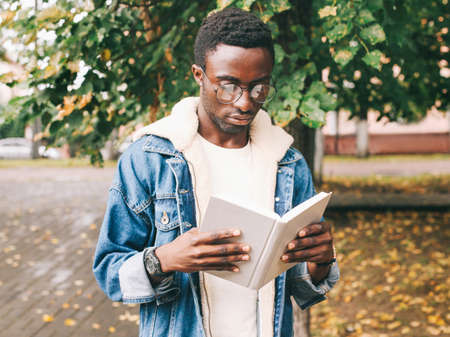 Portrait close up of young african man student reading a book wearing an eyeglasses in autumn city parkの写真素材