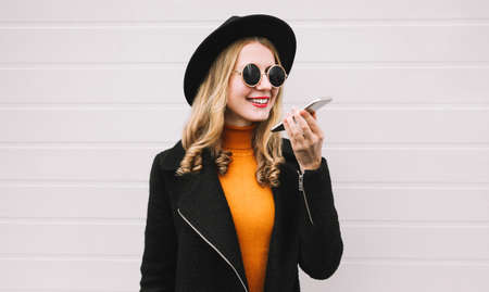 Portrait of happy smiling young woman with phone using voice command recorder or calling, wearing a black coat, round hat on gray backgroundの写真素材