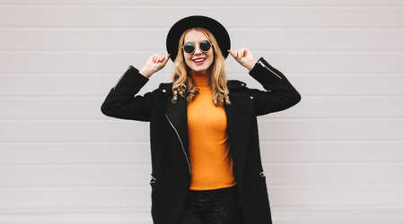 Fashionable portrait of stylish happy smiling young woman having fun female model posing wearing a black coat, round hat on city street on gray backgroundの写真素材