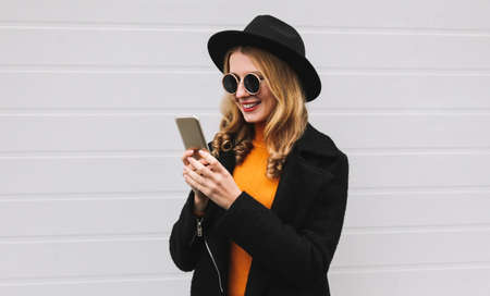 Portrait of stylish young woman model with smartphone wearing a black coat, round hat, sunglasses on gray backgroundの写真素材