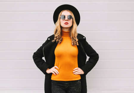 Portrait of fashionable young blonde woman model wearing a black coat, round hat on gray backgroundの写真素材