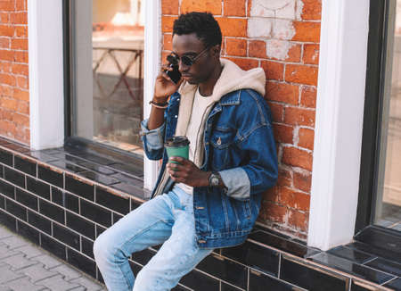 Portrait of young african man talking on smartphone while sitting with cup of coffee on a city streetの写真素材
