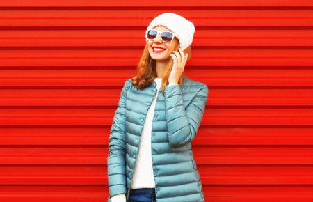 Portrait of happy smiling young woman talking on a smartphone wearing a jacket and white hat, jacket on red backgroundの写真素材