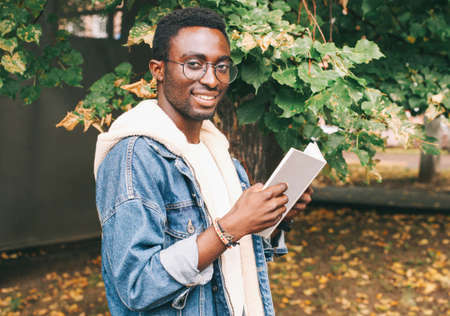 Portrait of young african man student with a book looking away wearing an eyeglasses in autumn city parkの写真素材